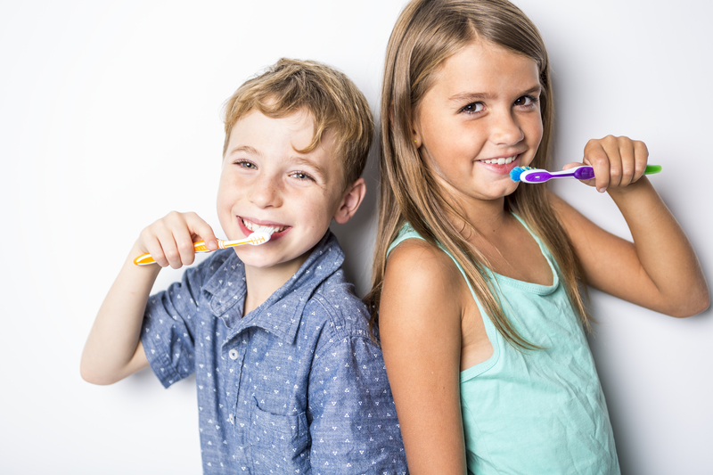 A young boy and girl looking at the camera, smiling and brushing their teeth.