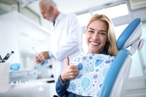 Woman sitting in a dental chair smiling, doing a thumbs-up while a dentist is in the background.