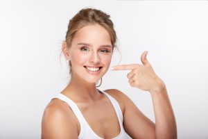 A blonde young adult woman that is smiling at the camera and pointing to her smile.