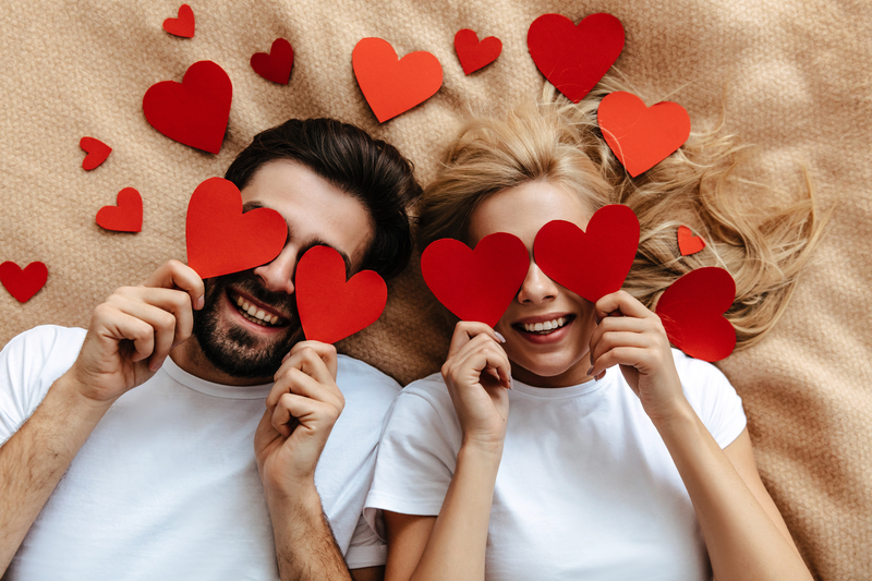 A couple lying next to each other surrounded by cut-out red hearts. Both of them are holding hearts over their eyes and smiling.