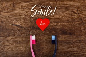 A wooden background that has the words "I love your smile" at the top and "love" in a heart cut-out below. There is a pink and blue toothbrush below that are facing one another.