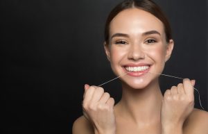 A beautiful, young adult brunette woman that is smiling at the camera and smiling as she flosses her teeth.