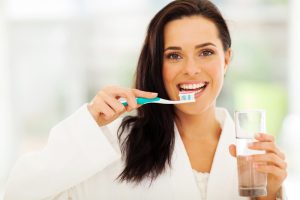 Woman that is brushing her teeth and holding a glass of water. 