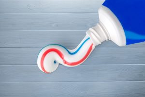 A blue tube of toothpaste that is squirting out onto a gray wooden backdrop. The toothpaste has a blue stripe and a red stripe in it.