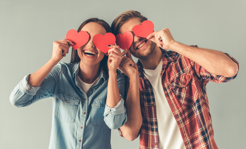 A couple holding red hearts in front of their eyes as their heads touch and they laugh.