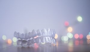 A light gray background with a few multi-colored lights. Sitting in front of the background is an Invisalign aligner.
